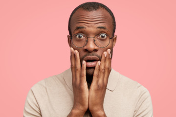 Photo of scared handsome dark skinned young male hears sudden negative news, keeps palms on cheeks,...
