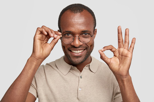 Dark skinned male with bristle and dark skin shows ok sign, has positive expression, wears spectacles, keeps hand on rim, has broad smile, stands over white background. Businessman approves something