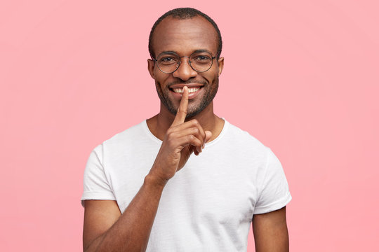 Positive dark skinned male makes hush gesture, has white teeth, asks to be quiet and not make noise, stands indoor alone, wears casual white t shirt. African American man shares his secrets.