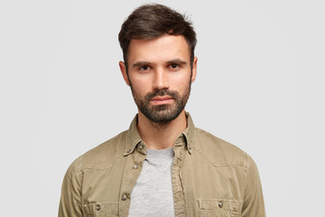 Obraz premium Photo of confident bearded male with dark hair, has serious facial expression, looks directly at camera, thinks about future job, dressed in fashionable shirt, stands against white studio wall