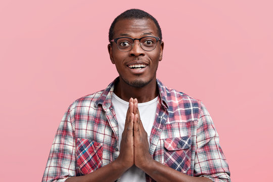 Attractive dark skinned young male student keeps hands in praying gesture, asks professor for good mark, dressed in checkered shirt, isolated over pink background. Please, help me, my friend
