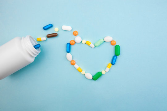 Multi-colored tablets and heart-shaped capsules, white bottle for tablets, pharmaceutical medicine pills on blue background, an analgesic against diseases