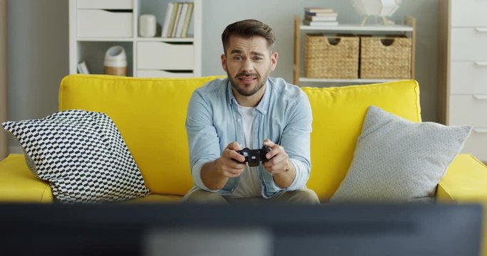 Caucasian young handsome man sitting on the yellow couch and playing video games, then winning. At home.