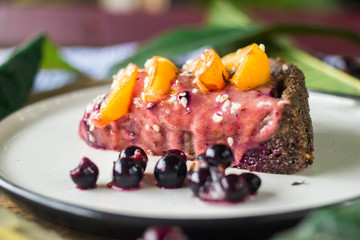 Piece of raw vegan pie with apricot and black currant berries cream. Vegan chocolate fruit cake with dates, banana, nuts, cocoa, carob powder. Vegetarian healthy dessert.