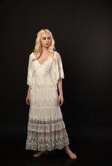 full length portrait of blonde girl wearing white lace dress. standing pose. black studio...