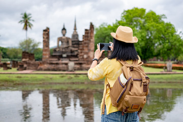 Obraz premium Asian tourist woman use mobile take a photo of ancient of pagoda temple thai architecture at Sukhothai Historical Park,Thailand. Female traveler in casual thai cloths style visiting city concept.