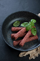 Frying pan with raw balkan cevapi or cevapcici sausages, vertical shot on a black stone background, selective focus