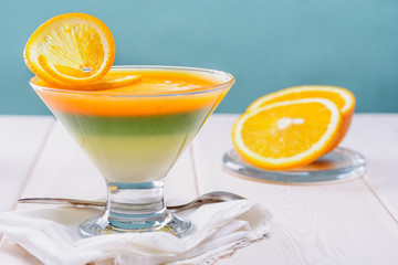 Colorful multi-layered jelly in a glass  is on a white wooden table next to slices of orange