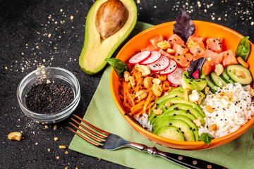 Hawaiian salad Poke with salmon fish, avocado, Korean carrots, cucumbers, radish and rice, with black sesame. in an orange plate. On the black background. copy space, top view
