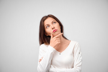 Close up portrait of pretty young woman thinking about problem, confused emotion, isolated, wearing white shirt, hipster style, dark hair, holding finger at lips, looking up, having idea