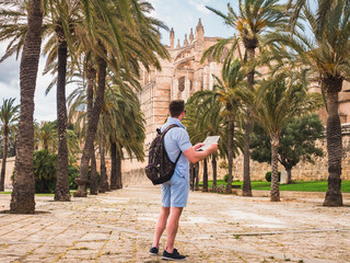 Attractive man with sunglasses and a backpack, looking at the map with sights on the background of palm trees and historical buildings. Spain, Palma de Mallorca © Svetlana