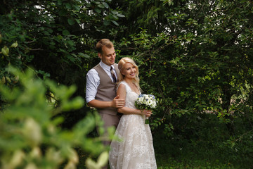 Happy newlyweds, Smiling stylish groom is embracing a charming bride, while standing behind her, at the wedding walk in the park.