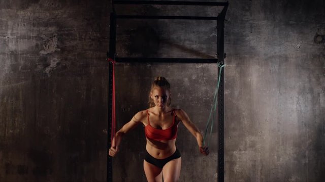 Strong young woman is doing physical exercises for developing muscles of hands, back and abdominal. She is stretching hard rubber ropes, fixed on a rack