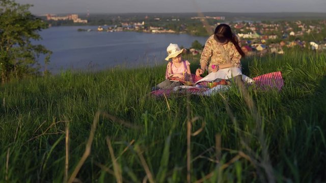 The family sits on a blanket on a picnic on a summer day on a hill overlooking a river or lake. Young mother and little daughter draw.