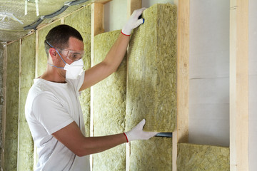 Worker in protective goggles and respirator insulating rock wool insulation in wooden frame for...