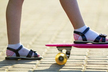 Child slim legs in white socks and black sandals on plastic pink skateboard on bright sunny summer...