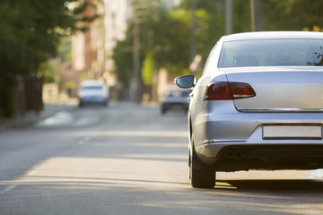 Close-up back view of new shiny expensive silver car moving along city street on blurred trees, cars and buildings background on sunny summer day. Comfortable transportation and speed in modern life.
