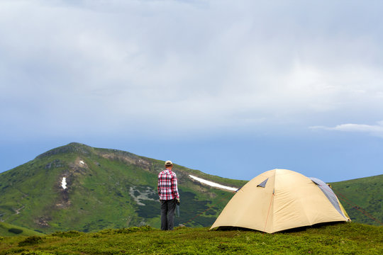 Young handsome man standing on green grassy valley on sunny day at small tourist tent mountains and bright blue sky background. Tourism, traveling, hiking and active lifestyle concept.