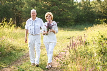 Naklejka na ściany i meble Elderly couple in the white linen dress walking outdoors, in the hands of a bunch of flowers, smiles on their faces, and on the background of coniferous forest