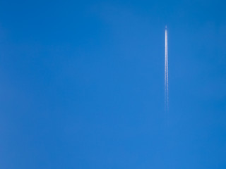 A jet airplane far away climbing fast with a long double white trail in a pretty clear blue sky, with a tree canopy in the bottom of the photo near the photographer