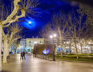Logrono, La Rioja, Spain. April 23, 2018: Nighttime image of a central plaza of logrono with the full moon partially covered by clouds, very illuminated by streetlights and people walking