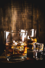 Two whiskey shot glasses on dark wooden background, with ice cubes, copy space