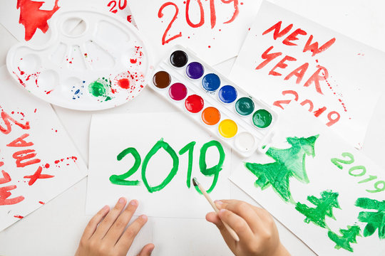 Children's drawings set by a watercolor on white paper of a congratulation by New year and Christmas. The child writes figure 2019 with largely green paint. A palette and a set of paints on a table. 
