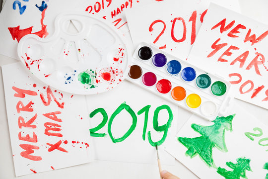 Children's drawings set by a watercolor on white paper of a congratulation by New year and Christmas. Figure 2019 largely green paint. A palette and a set of paints on a table. Horizontal shot