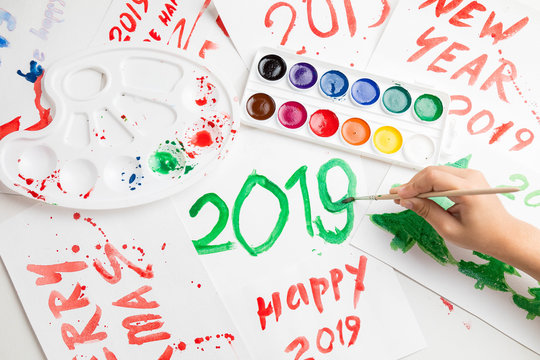 Children's drawings set by a watercolor on white paper of a congratulation by New year and Christmas. The child writes figure 2019 with largely green paint. A palette and a set of paints on a table. 