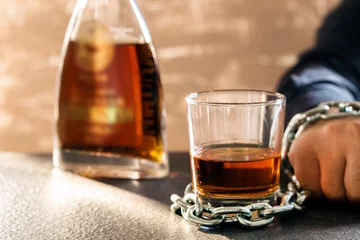 Rolgordijnen Bar De hand van de man is geketend aan een glas met alcohol en een fles cognac, cognac. Concept - alcoholisme en afhankelijkheid.