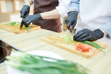 Closeup of unrecognizable female chef cutting vegetables standing at wooden table in restaurant kitchen with assistant, copy space