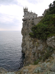 A castle with turrets located at the very edge of a cliff standing in the sea. With fences around the edges and tourists looking at local beauty from a bird's eye view.
