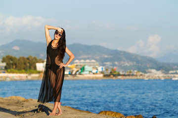 cute young woman with long dark hair in black swimwear and transparent dress on city and mountain background. concept of happy holiday and resort time