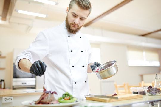 Portrait of handsome professional chef serving meat dish while cooking in modern restaurant kitchen