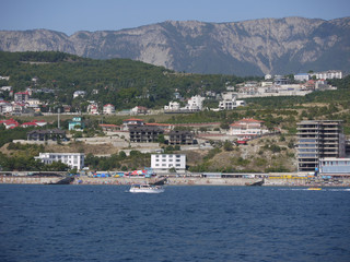 Fototapeta premium Boat sailing in the sea near the shore with tourists on holiday with the houses of the spa town seen on the coast against the backdrop of high mountains on the horizon.