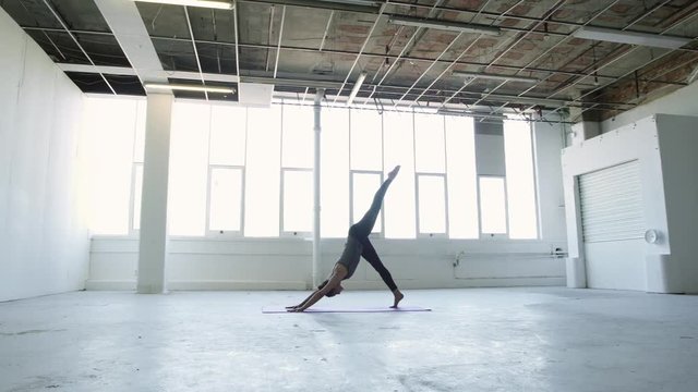 Wide orbit shot of a flexible young brunette woman practicing yoga moving from splits to a three legged dog in a white warehouse studio.