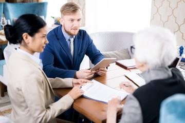 Young businessman with touchpad and his colleague with contract communicating with senior woman at the table at cafe