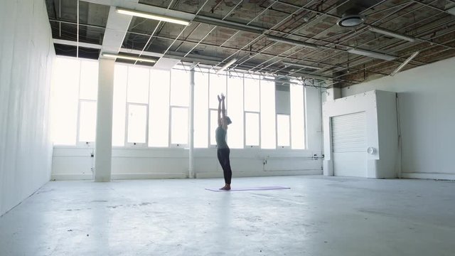 Wide moving shot of a young brunette woman practicing yoga doing a standing forward fold hamstring stretch in a white warehouse studio.