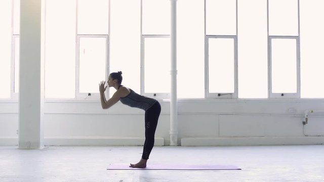 Tripod shot of a strong and young brunette woman practicing yoga sun salutation sequence in a white warehouse studio.