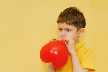 Child in yellow t-shirt inflates a red balloon on yellow background.