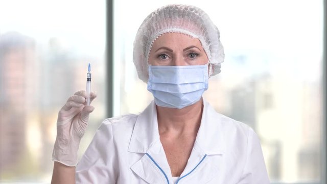 Middle aged doctor holding a syringe. Mature caucasian female surgeon with mask, hair cap, gloves, surgeon cloth indoor.
