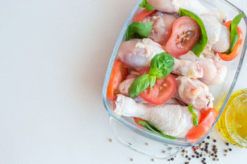 Raw uncooked chicken legs, drumsticks with tomatoes and basil in a glass backing dish, meat with ingredients for cooking, horizontal, top view, copy space