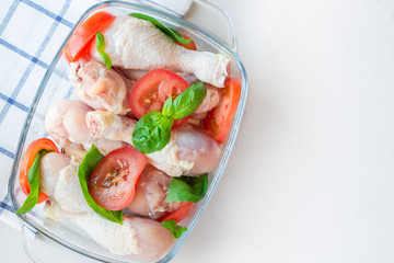 Raw uncooked chicken legs, drumsticks with tomatoes and basil in glass backing dish, meat with ingredients for cooking, horizontal, top view, copy space