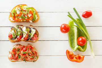 Bruschetta with fresh tomatoes and cheese, eggplant and mozzarella, sweet pepper and goat cheese on white boards with fresh vegetables. Top view. Copy space.