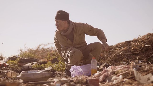 dirty homeless hungry man in a dump drinks the missing juice in the package with walking goes looking for food slow motion video. homeless dirty man roofless person looking for food in a dump. refugee