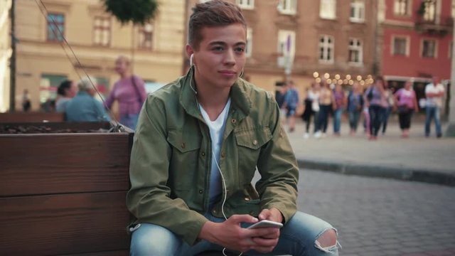 Handsome young man use phone listen music in headphone look around sitting on steet smile feel happy cell sunlight mobile gadget technology hipster close up slow motion portrait