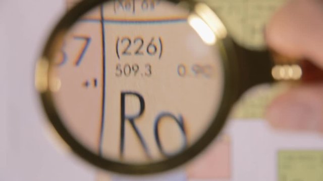 A hand moving a lens magnifier over a defocused board (a periodic table) and revealing the chemical element name, symbol and scientific properties of Ra, radium.
