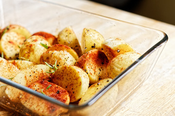 Preparing food - raw potatoes in rosemary, paprika and chili in glass dish on a wooden table prepared to be baked