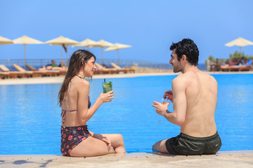 Beautiful young couple enjoying and drinking cocktails by the swimming pool on a hot summer day