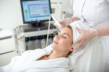 Side view beaming lady relaxing on couch while having procedure with special appliance in beauty salon. Health concept